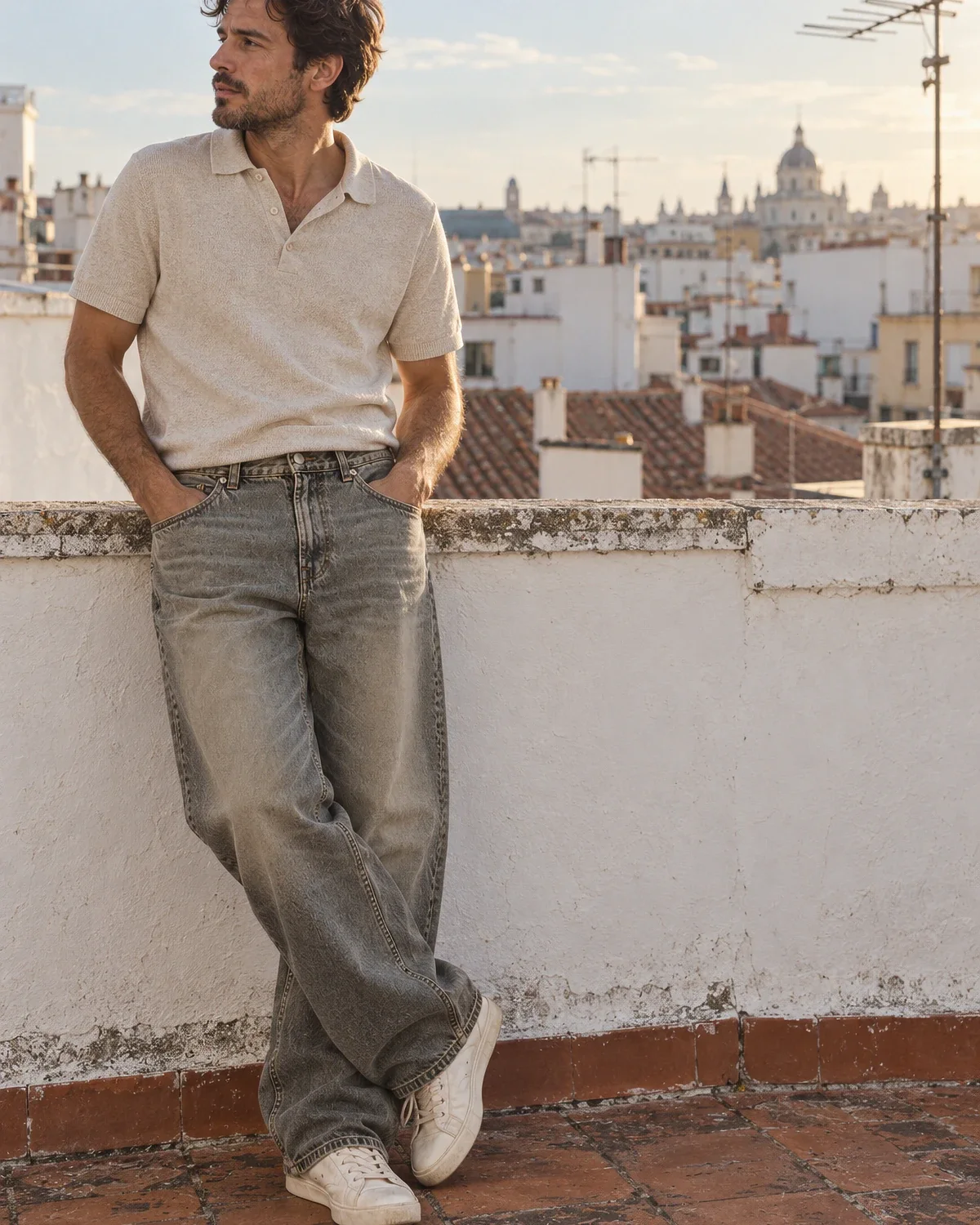 A man in olive-tinted baggy jeans and an oatmeal knit polo, leaning on a whitewashed rooftop wall in Madrid.